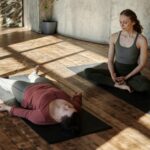 Two women practicing yoga indoors, one guiding the other in a relaxing pose on a wooden floor.